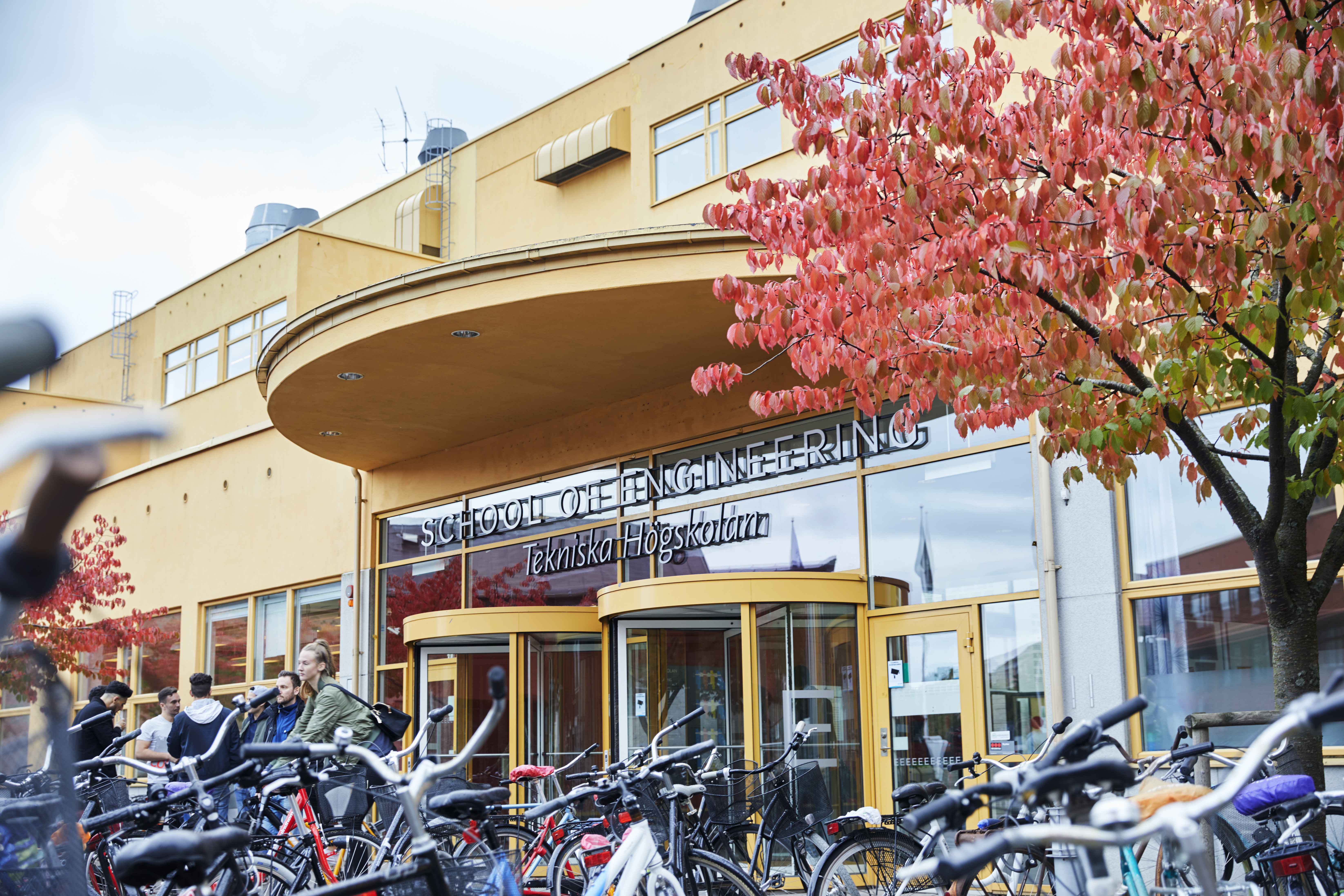 Entrén till Tekniska Högskolans gula byggnad med parkerade cyklar och några studenter i förgrunden.