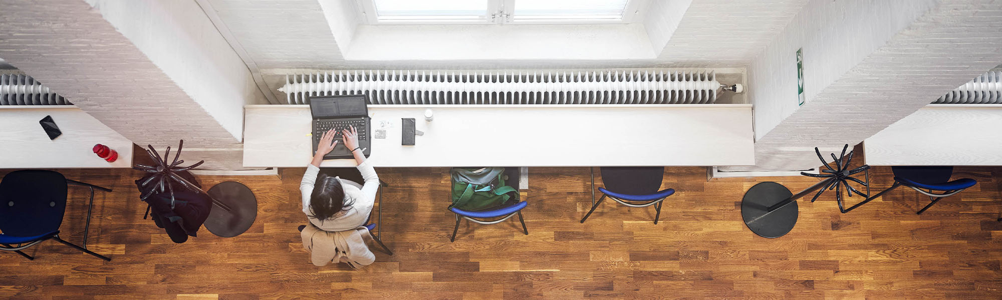 A student working on a laptop at a table next to a window. The picture is taken straight down from high up.