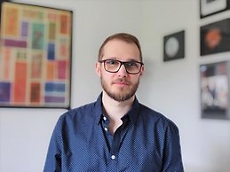 Portrait of a bearded man in glasses and a navy polka dot shirt standing indoors with modern framed art on the wall behind him.